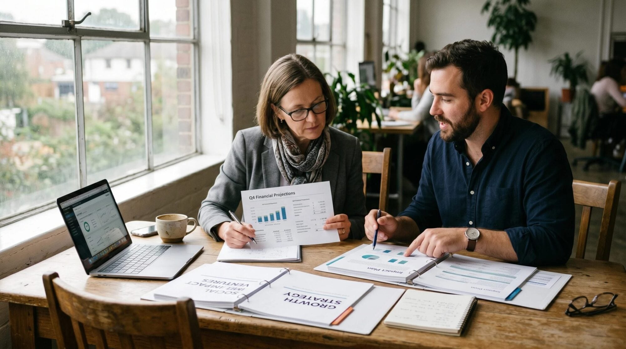 an investor and social entrepreneur reviewing financial documents and impact plans together at a wooden table in a natural light setting — papers, notebooks, and a laptop visible