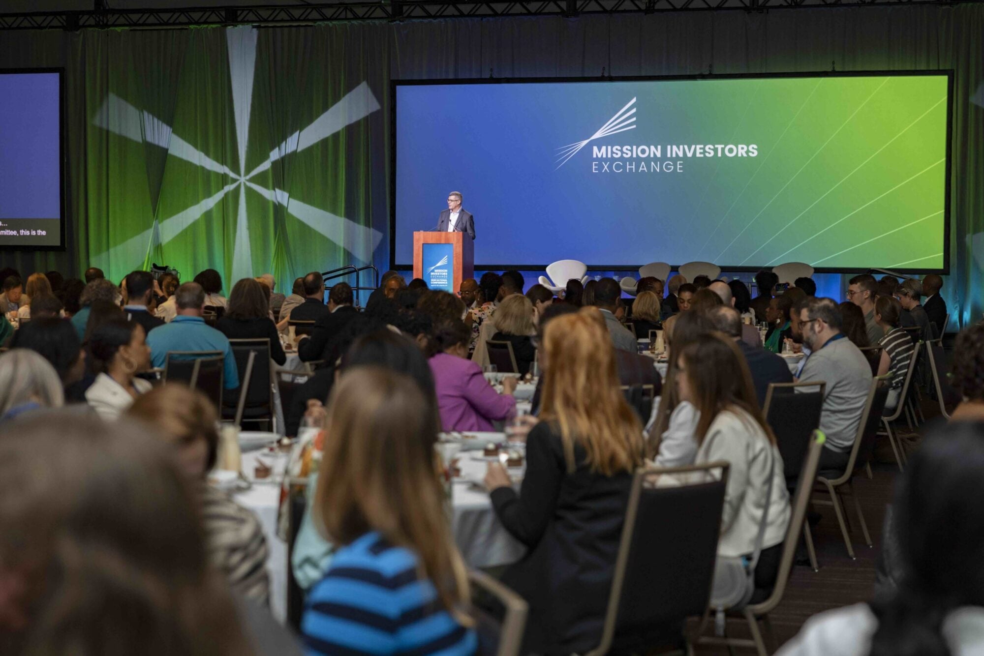 Speaker on stage at conference with attendees seated at tables