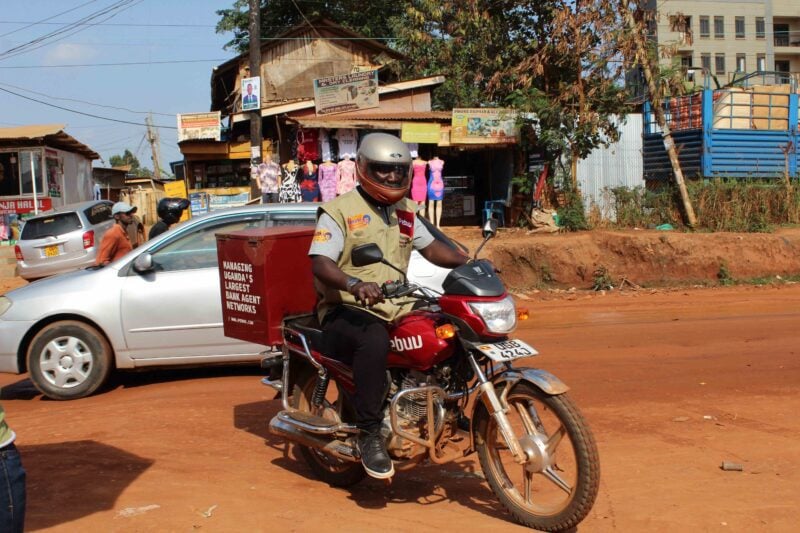 Helmeted Pebuu field team member riding a red motorcycle with a bank-agent network box on a busy unpaved street in Uganda.