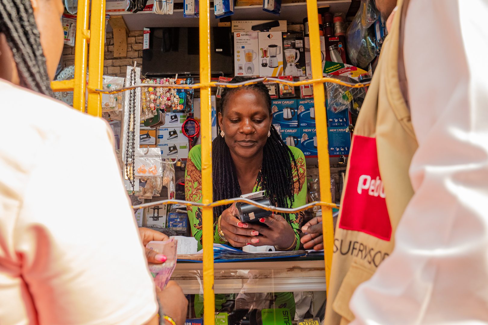 Woman completing a financial transaction at a small Pebuu-supported agent shop in Uganda, viewed through a yellow security grille, with customers in the foreground.