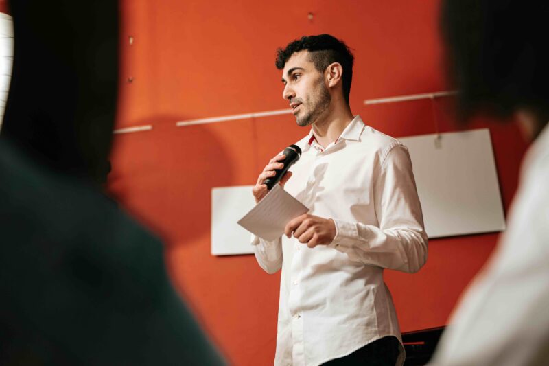 A man speaks into a handheld microphone while addressing an audience at an entrepreneurship or investment event.