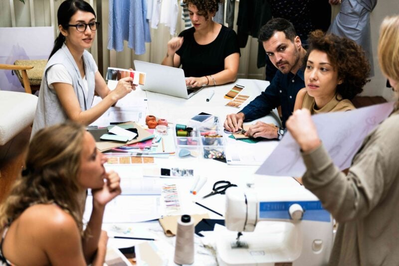A diverse team in a small fashion workshop reviews fabric samples and production plans around a table with laptops and sewing equipment.