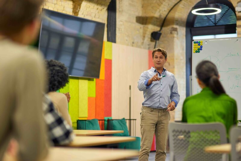 A man gives a presentation beside a whiteboard to a small group in a startup or innovation workspace.