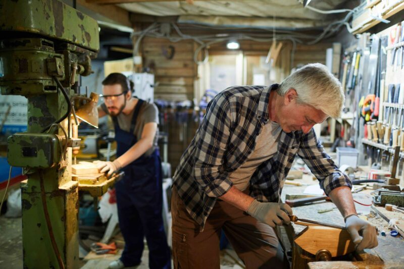 Older craftsman and younger worker working together in a well-used small business workshop.
