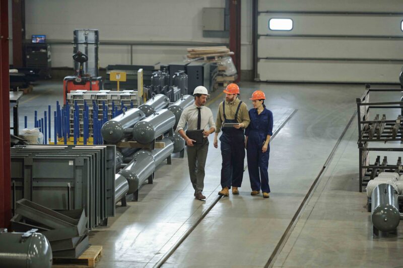 Three managers and workers walk through a large industrial facility surrounded by production equipment.
