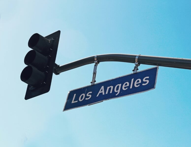A blue Los Angeles street sign hangs beneath a traffic signal against a clear blue sky.