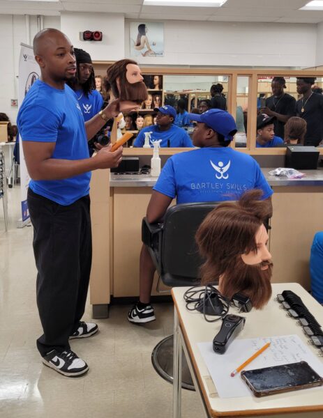 Chadwin Bartley holds a mannequin head and demonstrates barbering techniques to students gathered in a high school cosmetology classroom.