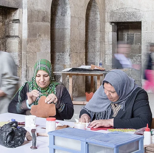 Two Mishkā artisans work on leather pieces at a table inside a historic stone building in Cairo.