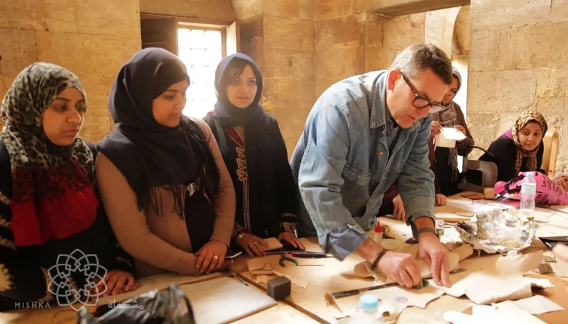 Bill Amberg demonstrates leatherworking techniques to a group of Mishkā artisans gathered around a worktable.