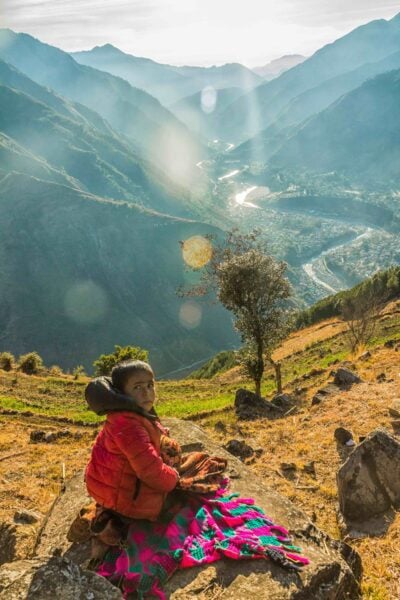 Child seated on a hillside overlooking a broad river valley and mountain community in Nepal.