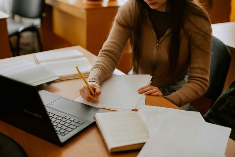 Student writing on paper beside an open laptop and several books on a library table.