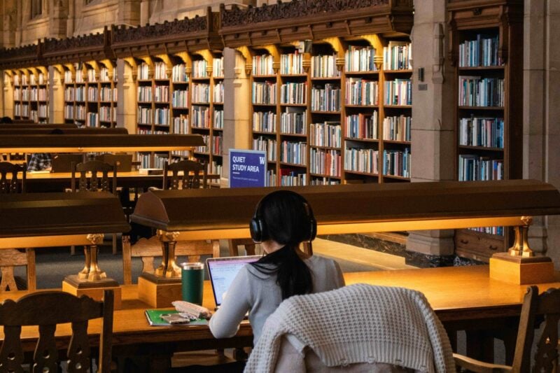 Person wearing headphones using a laptop at a wooden desk in a library reading room lined with bookshelves.
