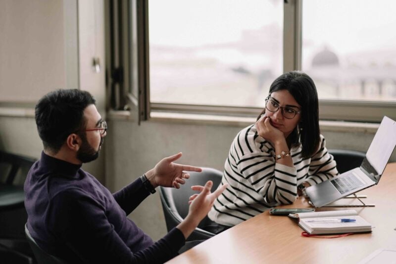 Two colleagues sit at a table by a window, one gesturing while the other listens beside an open laptop and notebook.