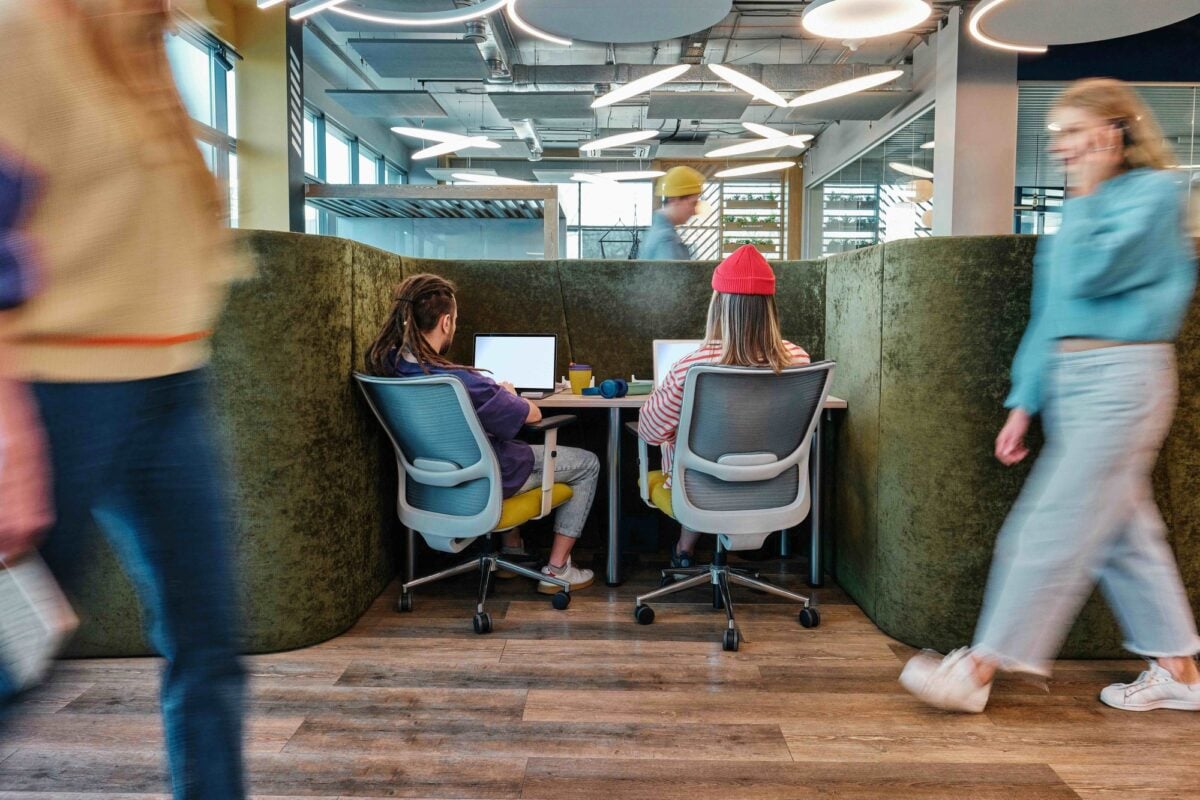 Two people work on laptops in a high-backed office booth as others walk past.