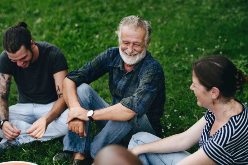 An older man and younger adults sit together on the grass, smiling and laughing.