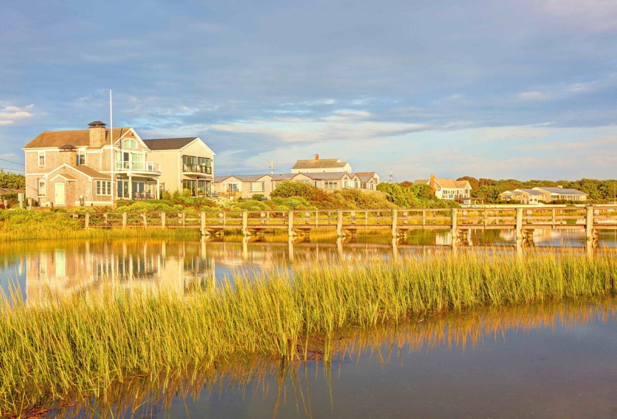 Coastal houses and a wooden walkway beside a marsh, with grasses and reflections in the water.
