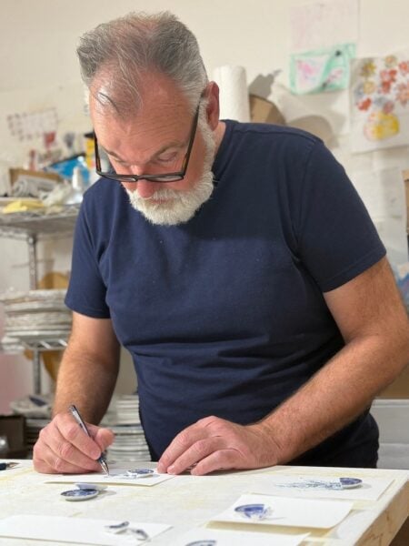 Rob Strati leans over a studio table, drawing beside small blue-and-white porcelain fragments.