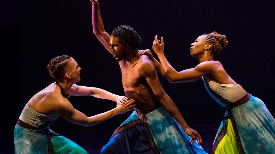Three dancers from Deeply Rooted Dance Theater perform on a dark stage, two reaching toward the dancer at center