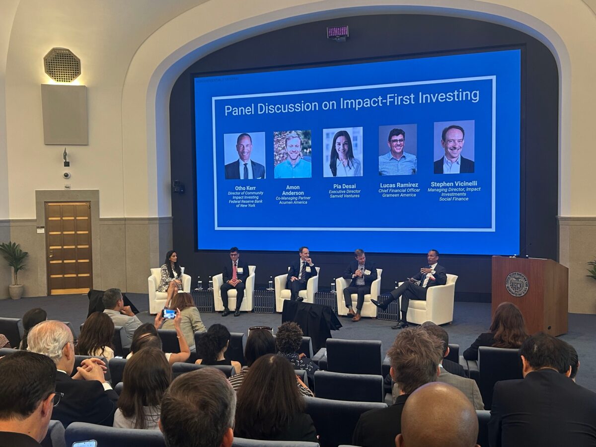 Five panelists sit on a stage at the Federal Reserve Bank of New York beneath a screen reading “Panel Discussion on Impact-First Investing,” with the audience in the foreground.