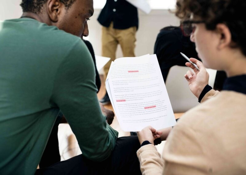 Two seated participants review a printed document with highlighted passages, one holding a pen beside the page.