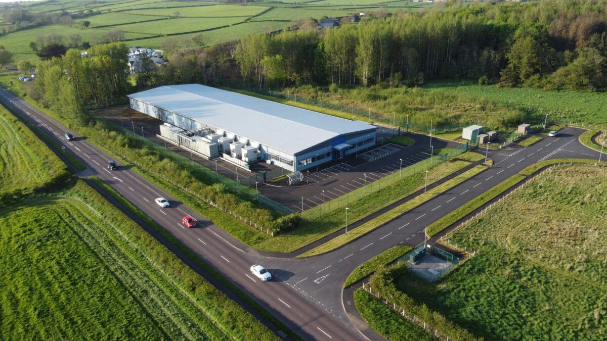 An aerial view of a long, white-roofed data center with a row of large equipment units along one side, beside a road with passing cars and green fields.