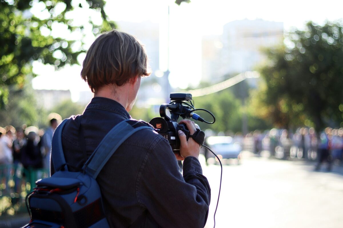 A videographer with a backpack, seen from behind, films a tree-lined street where a crowd has gathered.