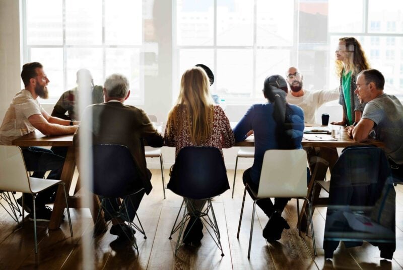 Nine people meet around a long wooden table in a bright office, with papers, a coffee cup and water glasses on the table.
