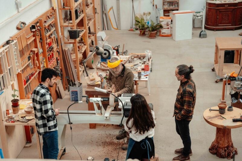 Four people gather around a wood lathe in a workshop lined with hand tools and woodworking equipment.