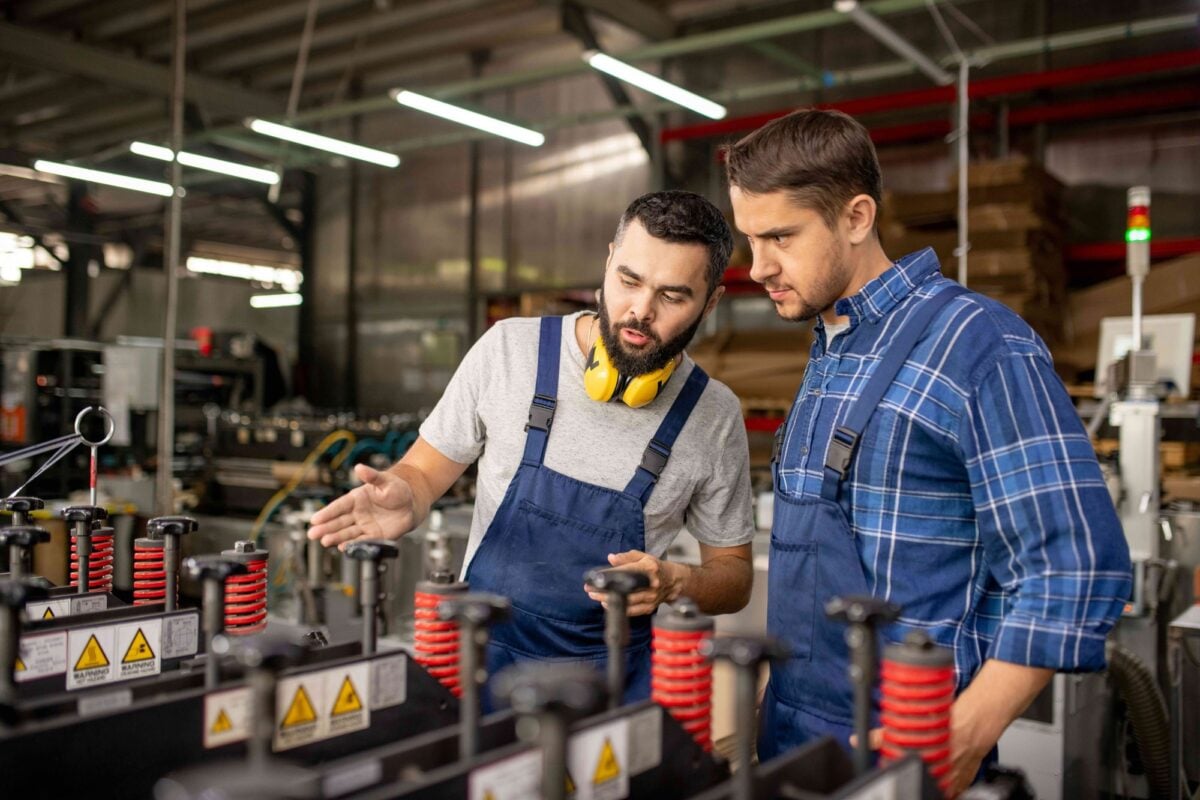 Two workers in overalls examine machinery, with one gesturing toward the equipment.